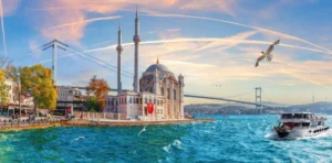 Panoramic view of Ortaköy Mosque and Bosphorus Bridge in Istanbul at sunset, with a ferry and seagulls over the Bosphorus.